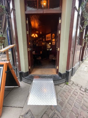 Metal ramp placed at step before entrance. Double wood-panelled glass doors immediately after ramp. Push brass plates at centre to open.