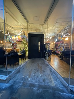 From sidewalk facing venue. Path to entrance ahead. Glass walls either side.