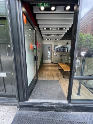 Single glass door with black frame. Push black handle on right to open. Loose textured doormat in front of door.