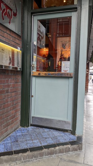 From street looking straight at main entrance at corner of venue. Single standard step up to doorway. Regular manual door opening inward with push handle on left side. Ramp available upon request.