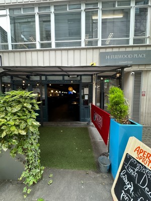 From pavement looking straight at opening ahead. Planters to left and right. A-frame on right obstructing path. Grass path ahead.