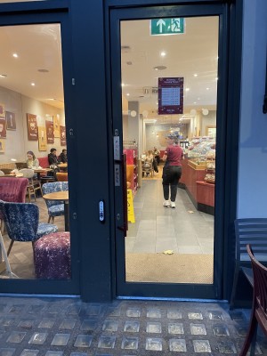 View of main entrance from street. Glass wide door which pulls opens outwards. There is a press for assistance button available to the left of the door