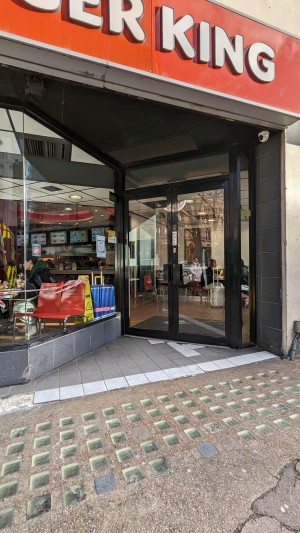 From street to right of venue looking left at alternative accessible entrance. Double manual glass doors opening outward. Back seating area upon entry, service counter to left.
