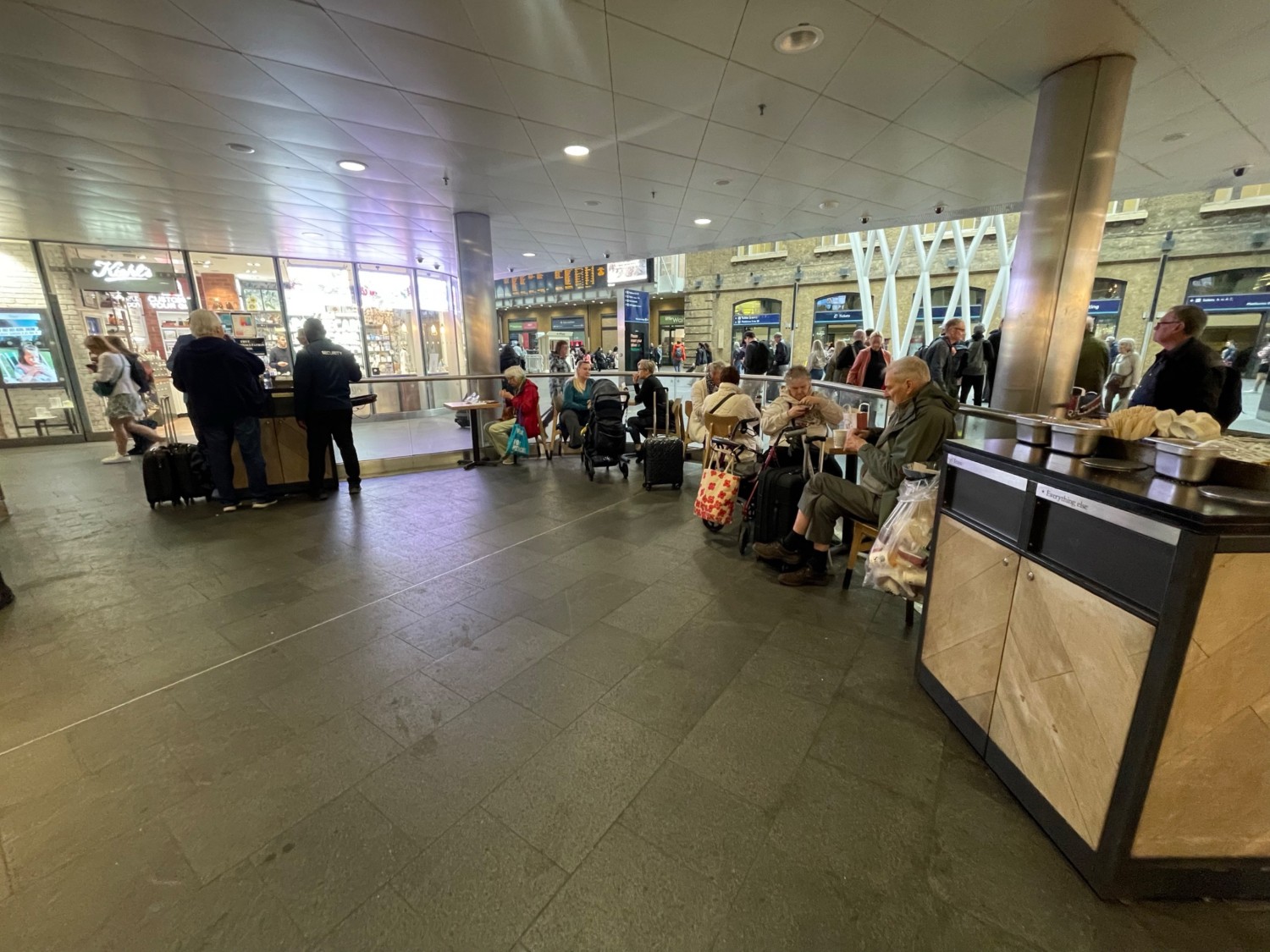 From entrance looking right. Widen area ahead. Coffee station on right. Small tables with chairs behind along curved fence on right and back.
