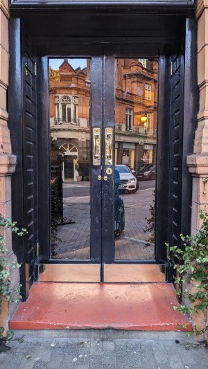 From street looking straight at main entrance. Single standard step up to double manual glass doors opening outward.