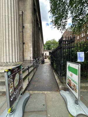 From pavement looking straight at built-in ramp ahead with black handrail along left. Standing signs to left and right.