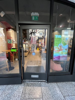 Glass door with black frame ahead. Pull handle from left to open. Doormat following.