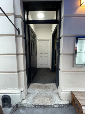 From sidewalk facing venue. First door ahead. Black wooden door. Push on right to open.