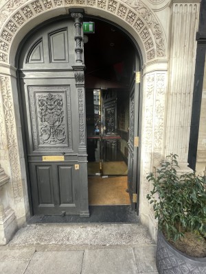 View of main entrance from street. There are double doors which push open from the middle. Beyond that there is another set of double doors which pull open.