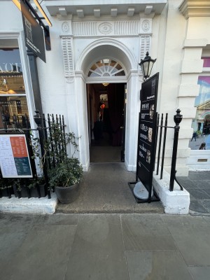 View of the front entrance to the building. There is a step which leads up the door. The door is a single door which pushes open from the left.