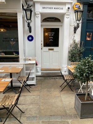 From sidewalk facing venue. Path ahead to entrance on back. Planter on right, outdoor seating behind. Outdoor seating along left.