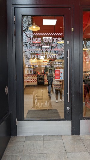 From street looking straight at main entrance. Wide glass manual door opening outward with pull handle on right side.