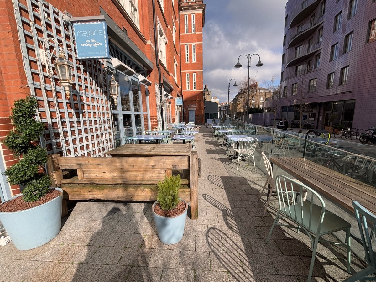 From A sign facing straight. Bench at left. Tables and chairs at its back. Tables and chairs along right.