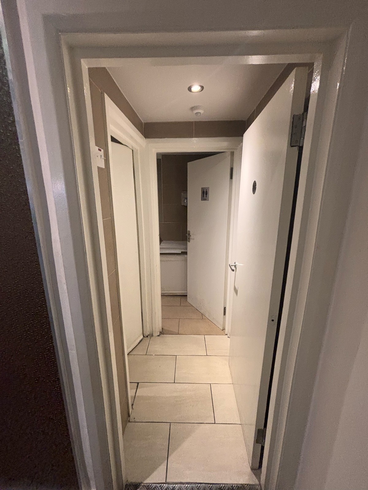 From end of path facing left. White bathroom door ahead. Push metal handle at center left to open.