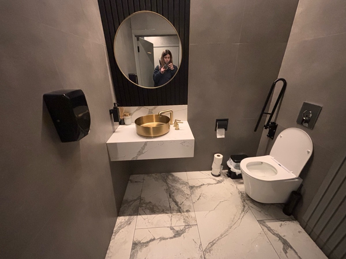 From bathroom door facing straight. Sink at back left. Toilet at back right. Grab rail at its left.