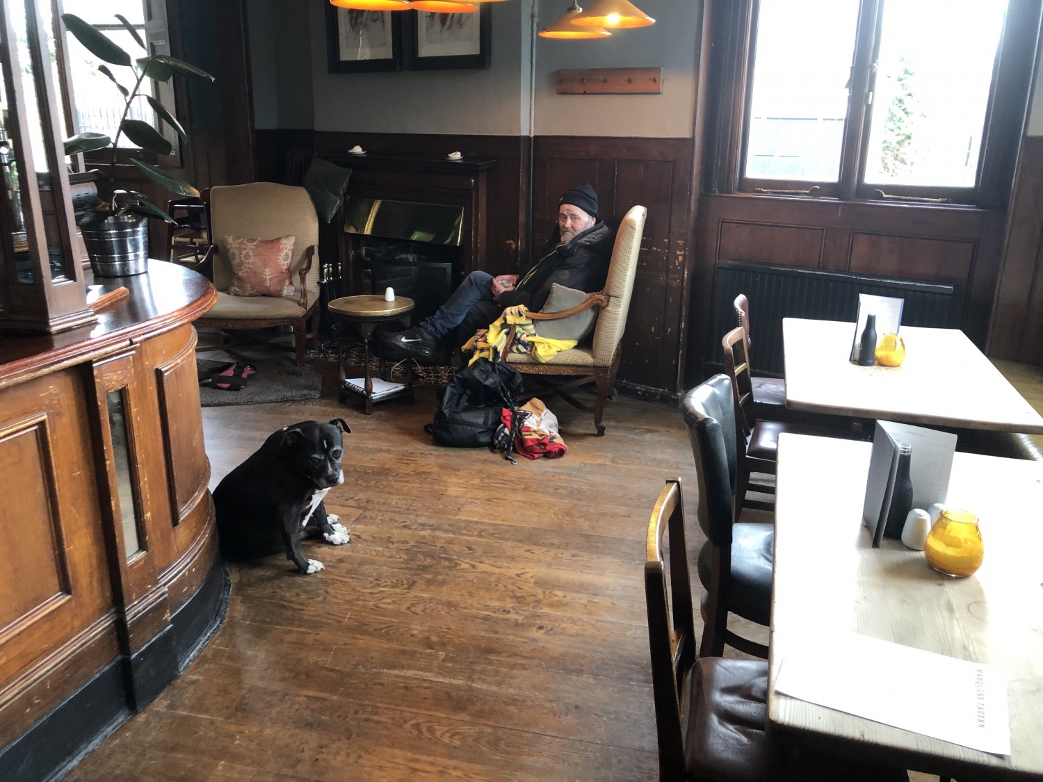 Photo of indoor area from entrance looking right. Bar to the left, seated height tables and chairs along right wall. Ahead is a low table with two armchairs.