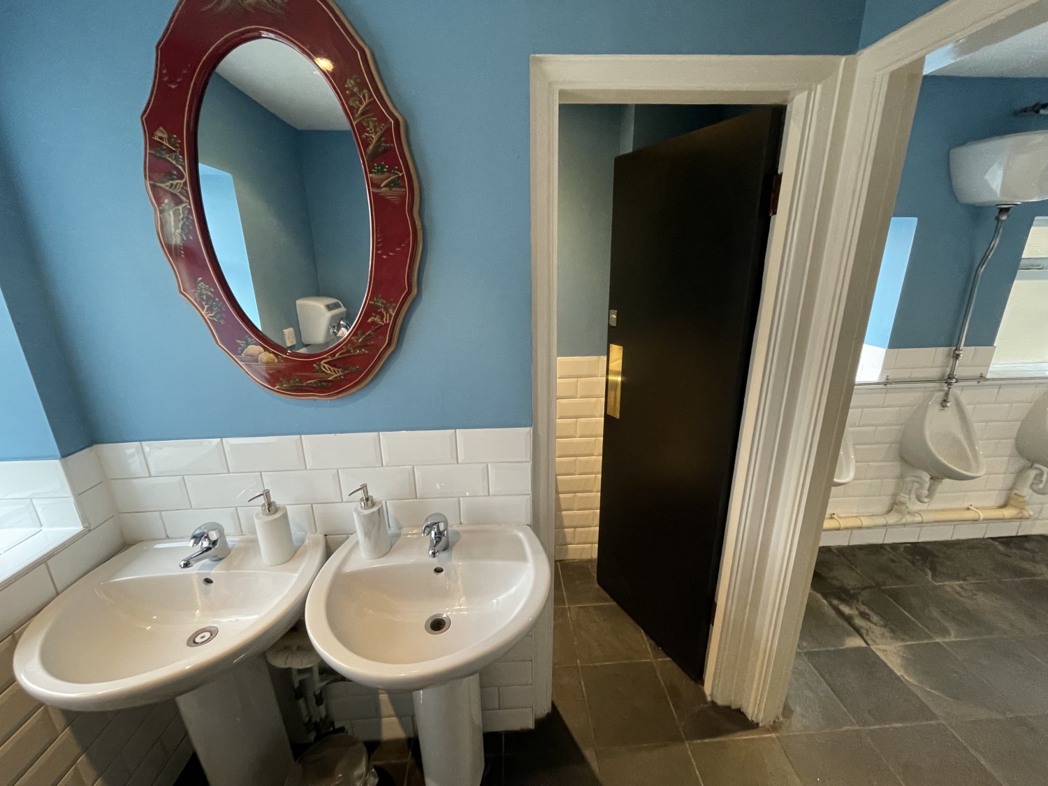 Front view upon entering gents bathroom. To the left there are two obstructed underneath sinks. Directly in front of entrance is a cubicle with a push door. Should be pushed from the left.