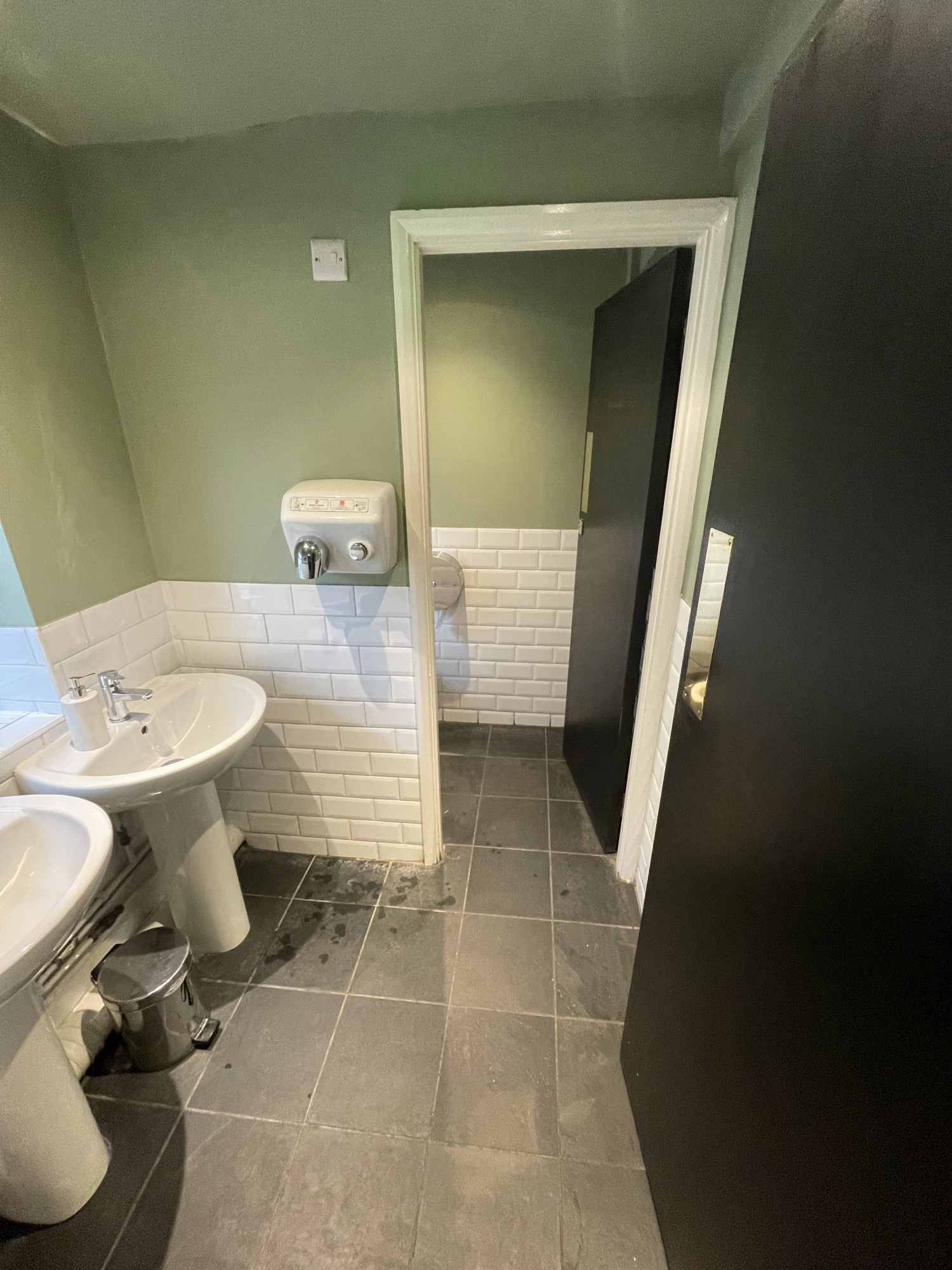 View from turning right inside ladies. There is a preceding door that is a push door to be pushed from the left leading to obstructed underneath sinks by the left wall. There is a hand dryer on the wall opposite when entering. There is then another left push door that leads to a cubicle in front.