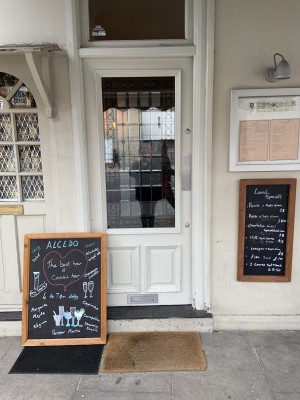 The door is on the left hand side of the restaurants. It pushes to open. There is a sign slightly obstructing the left hand side of the door. There is a door mat outside the door.