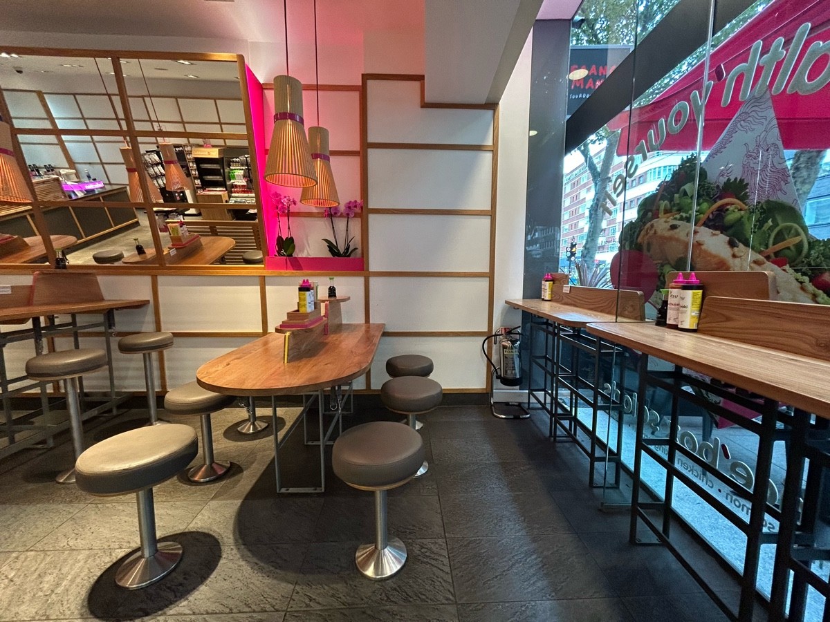 From end of order screen facing straight. High tables along right wall. Rows of tables along back wall with paths in between each.