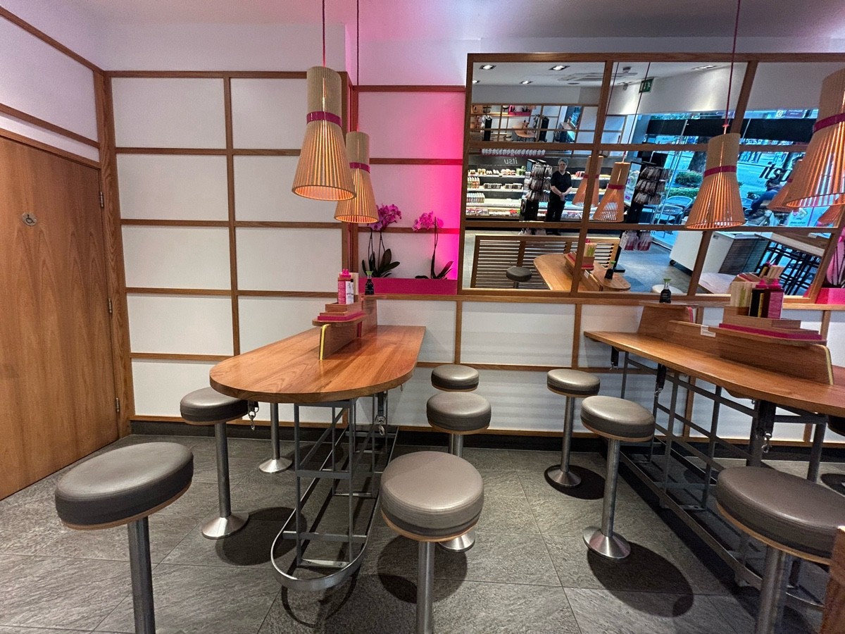 From end of order screens facing right. Tables and stools ahead on left and right. Paths between tables. Toilet on back left.