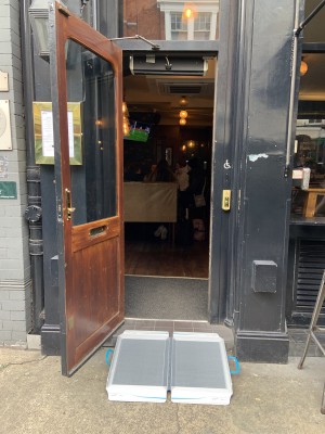 Black and glass panelled door. Gold vertical handle on right, pull to open. Ramp available upon request laid out leading up to door.