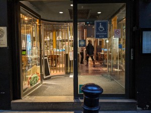 Glass doors with black frame and metal handles in centre. Push to open. Doorbell right of doors.