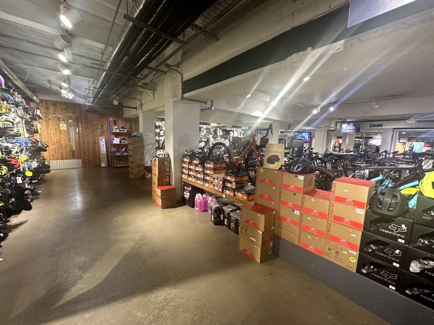 View of the downstairs area. After the wall with the bikes on display, turning left, there is a pathway with a shelve of equipment on the left hand side. On the right hand side there is a shelve with helmets on display.