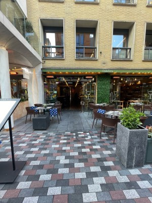 From courtyard facing venue. Path to entrance ahead. Menu sign in front on left.  Planters on right. Seating either side along path.