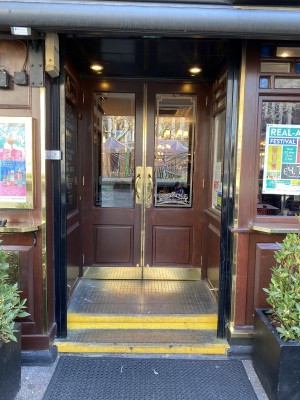 Directly facing the entrance of The Moon Under Water pub. The entrance has double doors with door handles located in the middle of the doors. There are two steps to go into the venue.