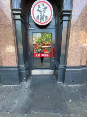 Single glass door with grey frame. Pull red vertical handle on right to open. Path leading to door slightly sloped up.