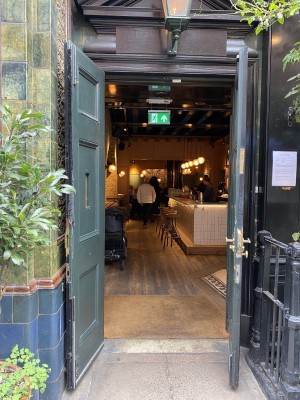Directly facing the entrance into the restaurant. On left is a big pot plant. In middle, double doors have been propped open outwards (door handle to left side of right door). On right is a black fence.