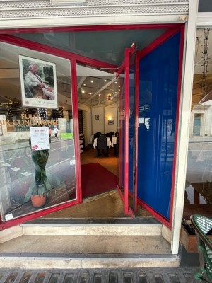 Glass door with red doorframe to far left of angled wall ahead. Vertical red handle to left of door, pull to open.