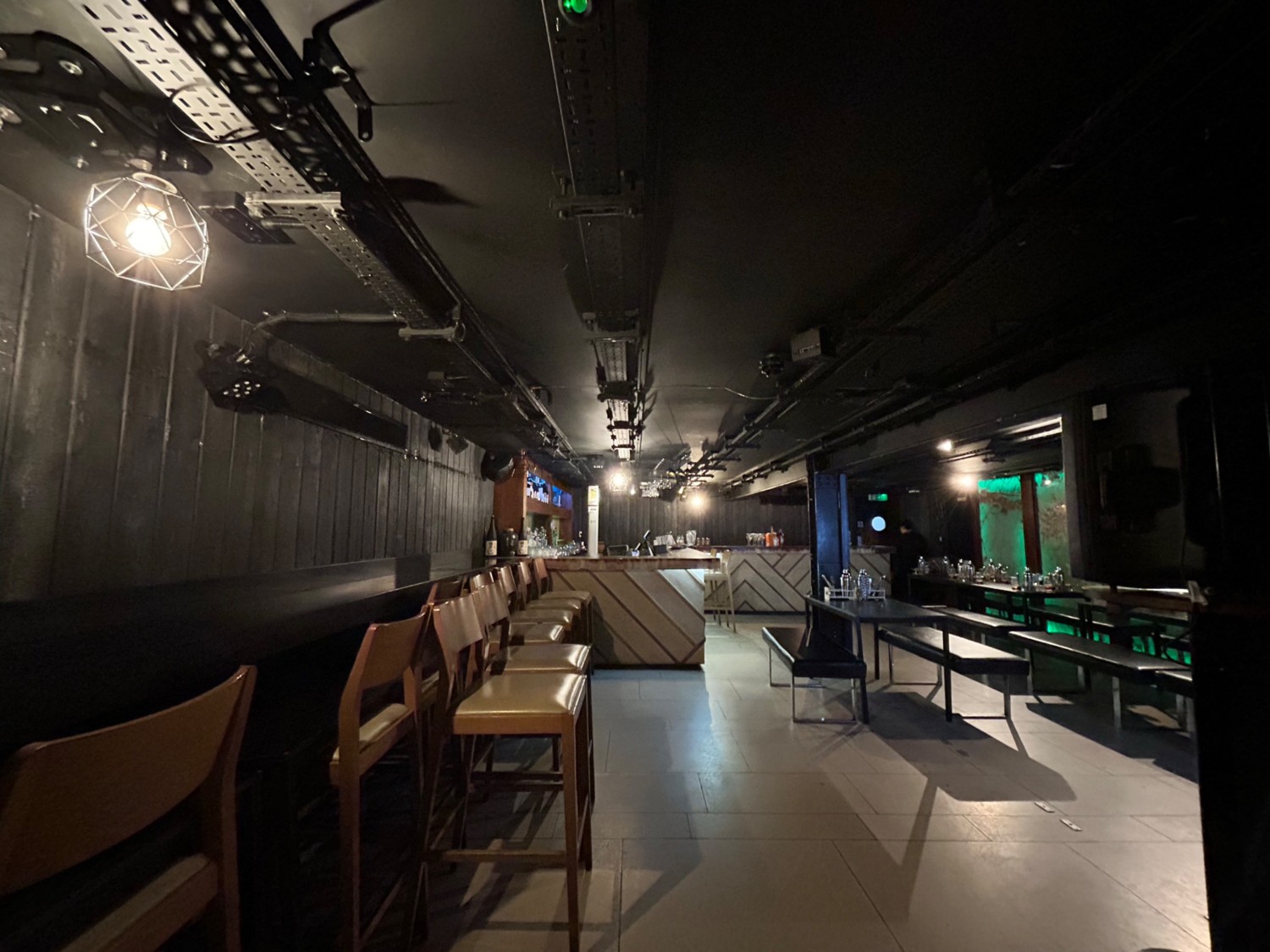 From doorway, facing straight. High tables and backed stools on left. Tables and benches on right. Path in middle leads to bar on back left.