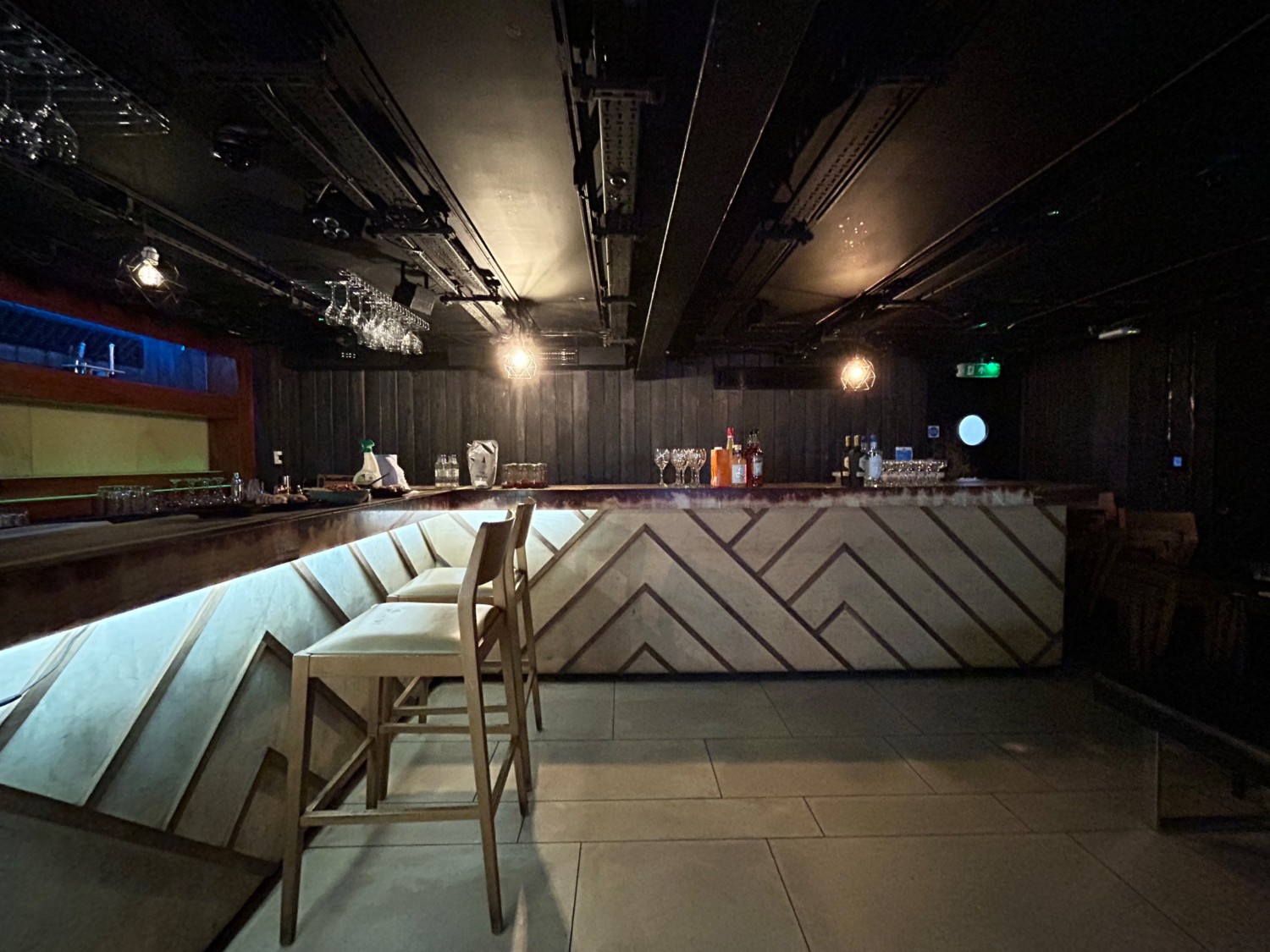 From middle of benches, facing straight. Bar along left wall and back, lined with backed stools. Path ahead.
