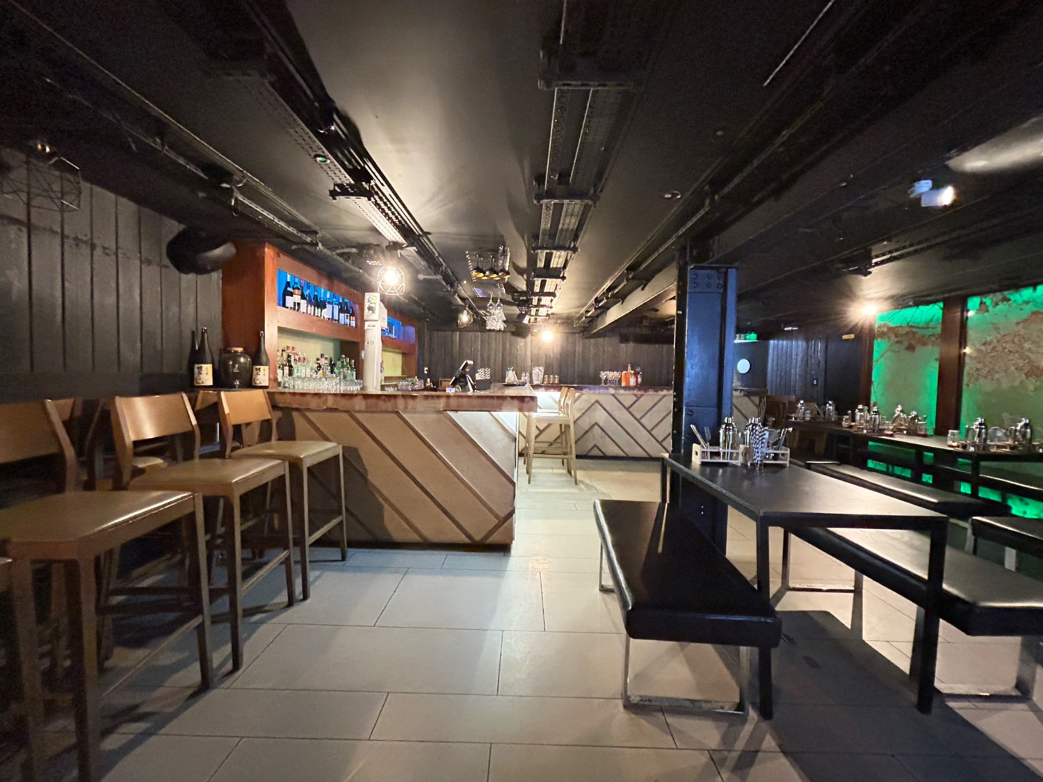 From middle of high tables, facing straight. High tables and stools along left, then bar beyond. Tables and benches on right.