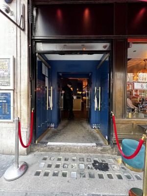 Glass double doors. Long vertical handle in the middle of both doors. Push to open. Second set of doors following. Pull handles in centre to open.
