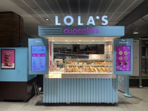 View of the venue which is a stand within London Bridge, by the tube station. There is a high counter, with a glass display showcasing cupcakes and cookies on offer. On either side of the counter are signs showing a menu.