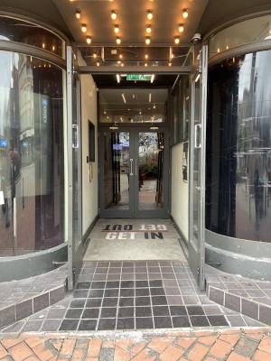 (1) Entrance to left of venue. Uneven built in ramp before first doors. Ground between first and second doors slopes up slightly. Both sets of doors pull to open by high hand bars where doors meet.