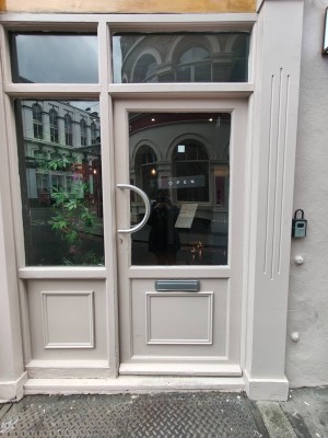 Facing pink framed entrance door. Push curved silver metal handle on left to open.