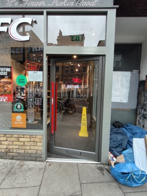 Facing grey framed glass door. Push red metal handle on left to open.