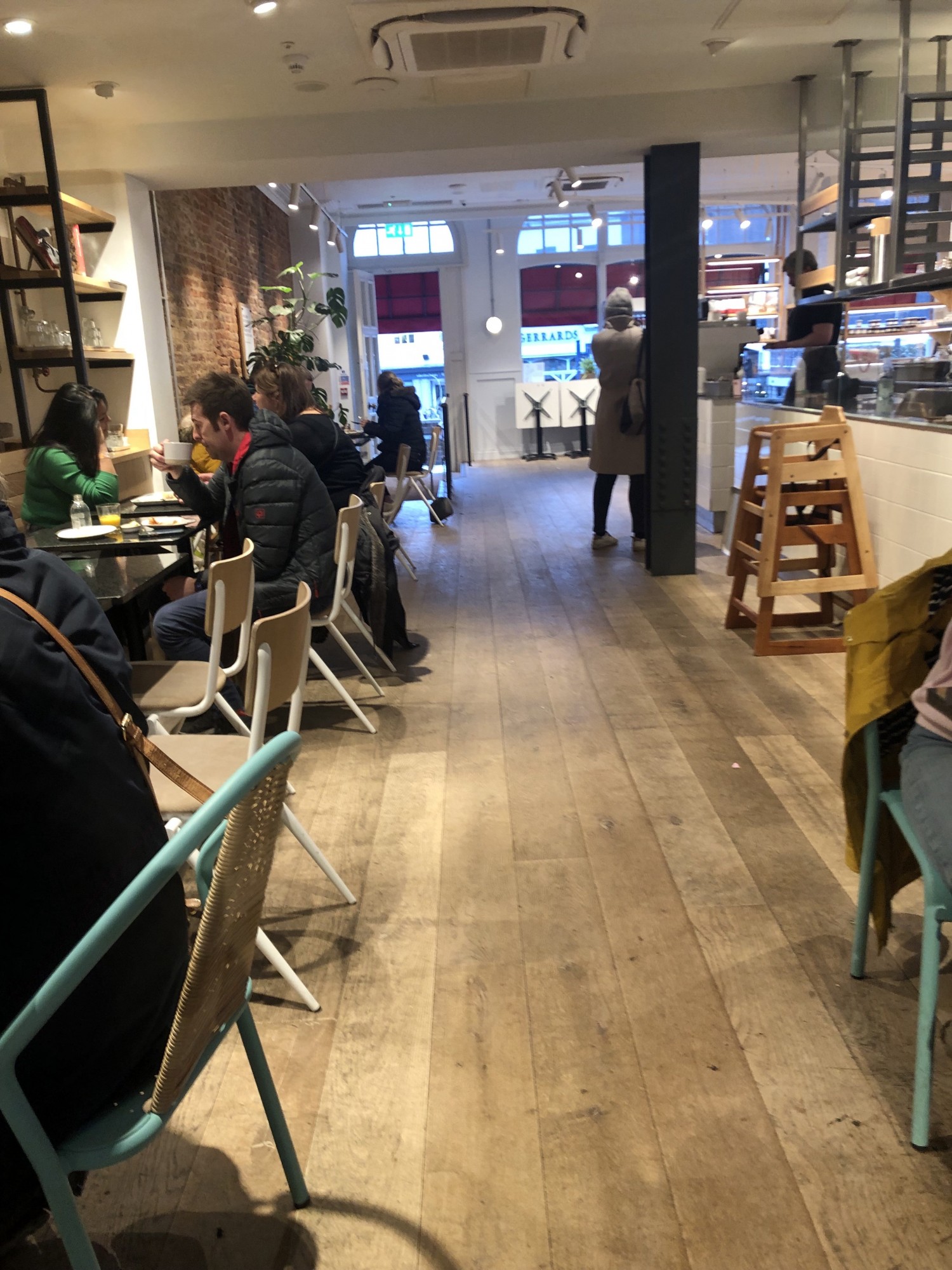 Cafe seating with customers on left (looking from back of cafe towards entrance). Stack of high chairs and counters on right. Wide open pathway between the two with wooden flooring.