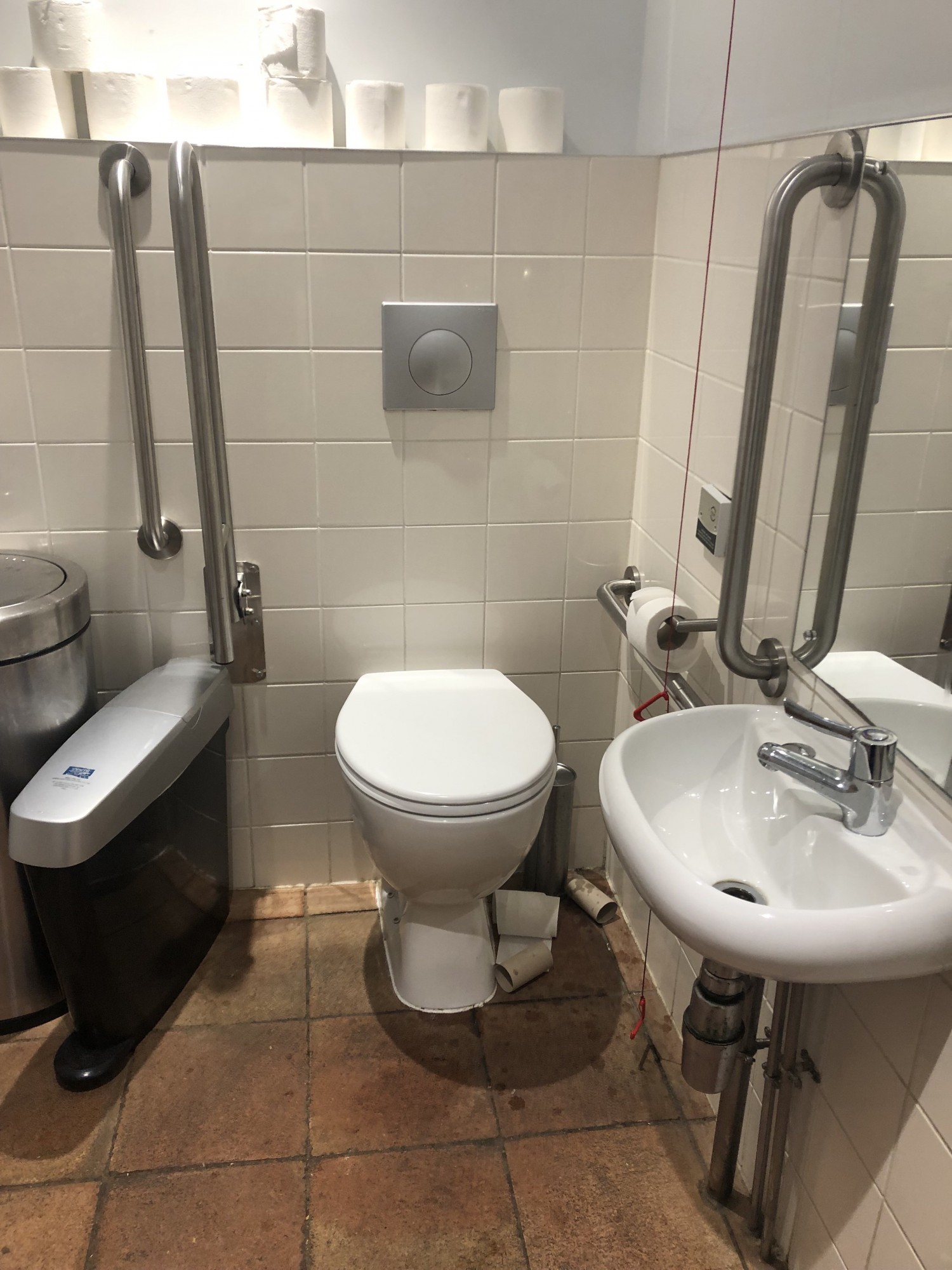 White bathroom with lid down, emergency alarm hanging freely, one fixed and one fold down grab bar to the left of the bathroom as you look at it. Sanitary bin and rubbish bin are pushed close to bathroom, also to the left. To the right, looking T bathroom, is bathroom roll and a horizontal fixed gra