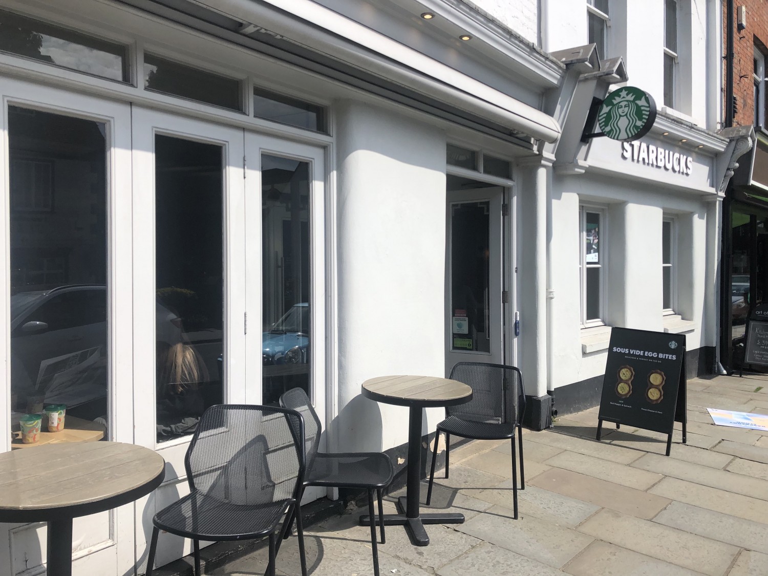 Exterior of Starbucks with several angles and chair sets, white wood frontage and glass windows. Doorway is below green Starbucks sign.