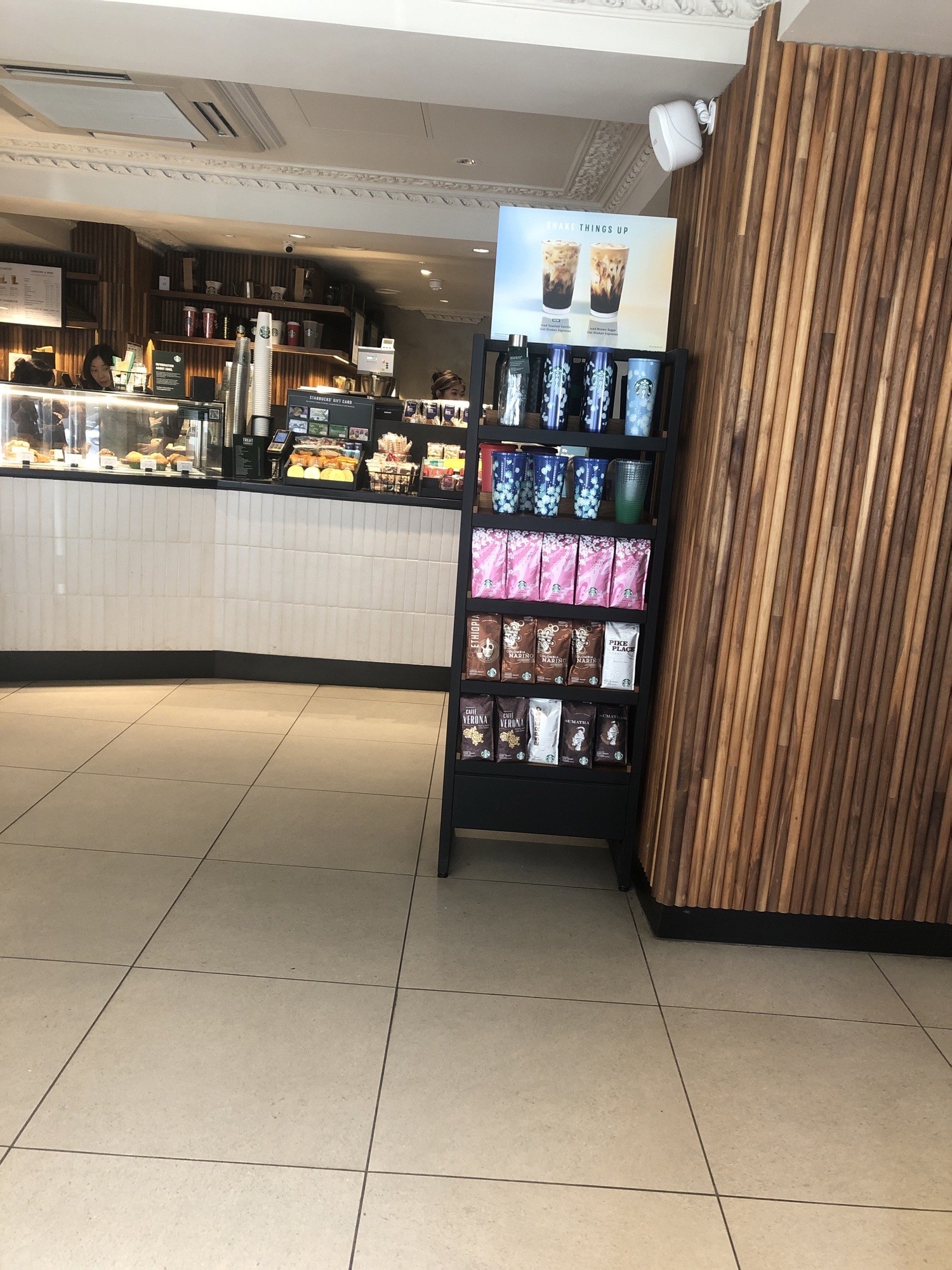 Main serving area at front of Starbucks with shelf display on right. Serving counter is white tiles in front and black contrast surface and glass around food. Floor is large light grey tiles with dark grout making square pattern.
