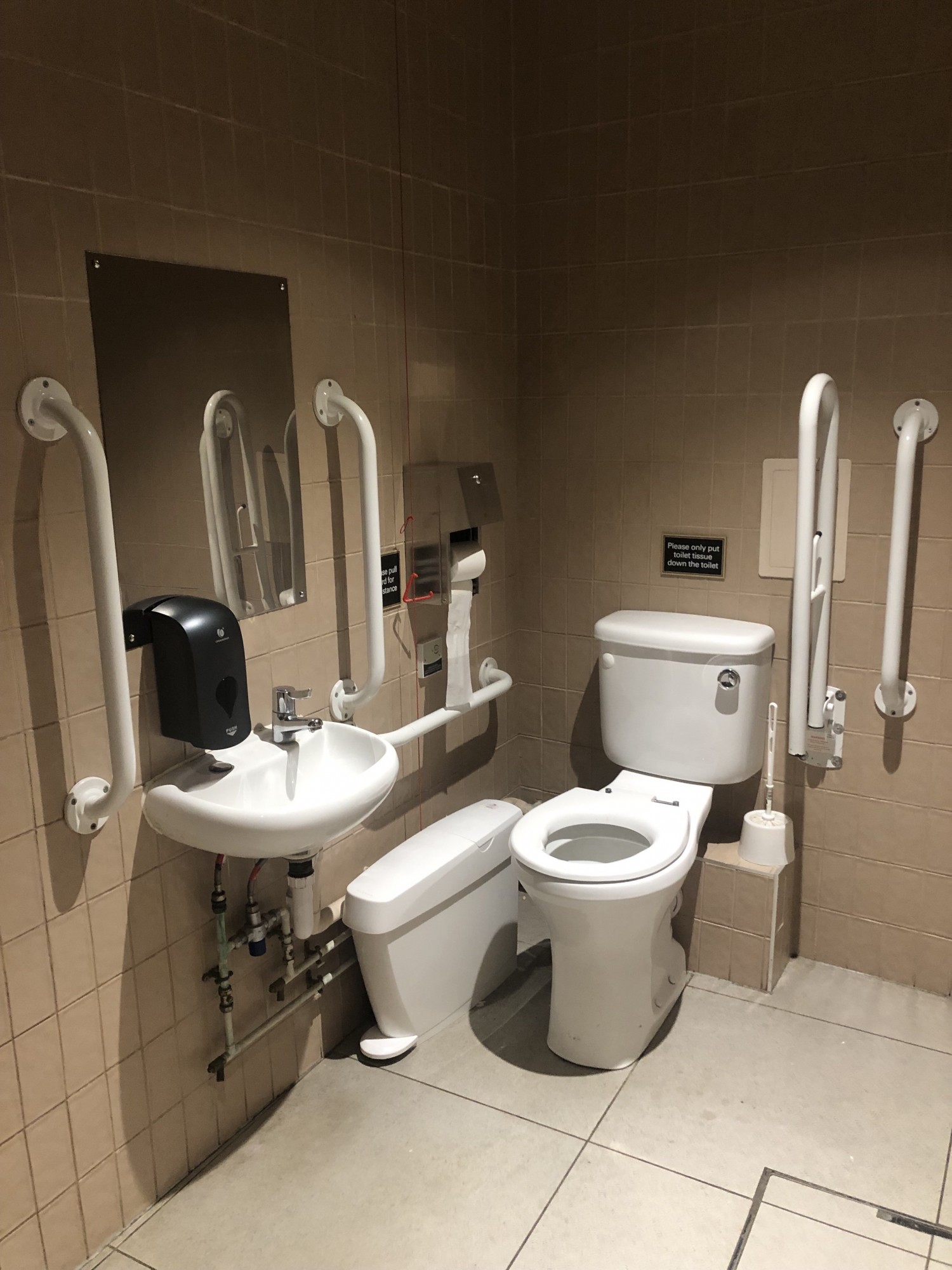 Right transfer white bathroom with white seat and white sink. The grab rails are also white and horizontal fixed and drop down either side of bathroom as well as fixed vertical either side of sink. Beige walls and white sanitary bin between bathroom and wall. Emergency cord hangs behind sanitary bin