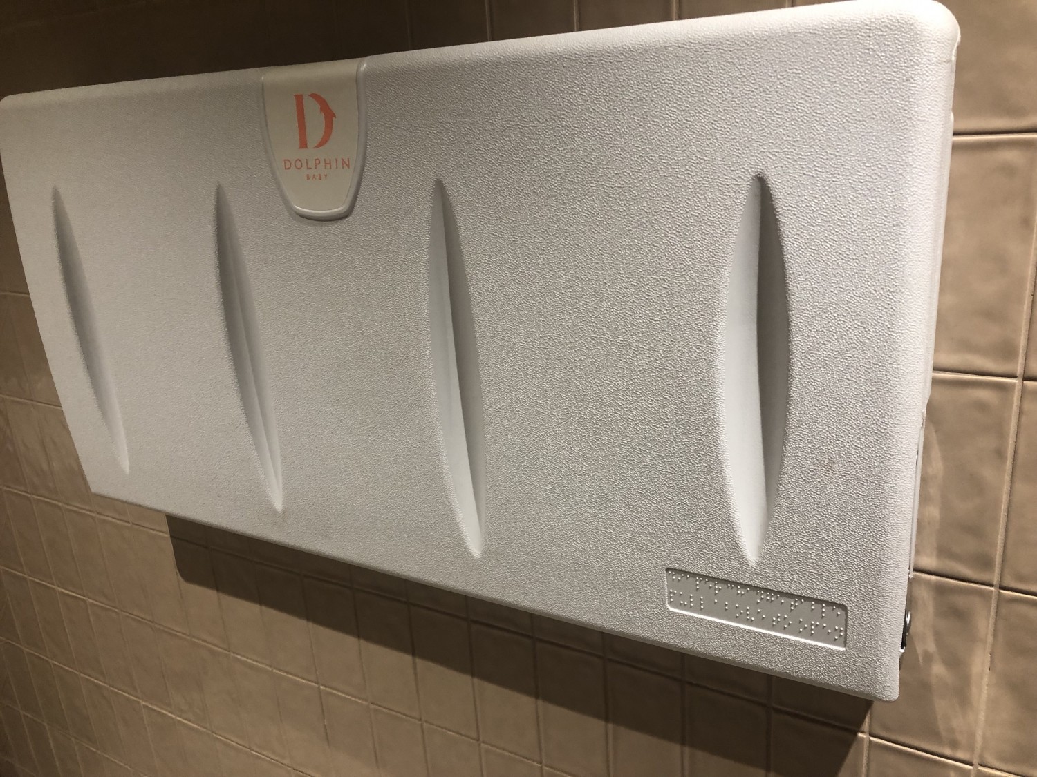 Off white fold down plastic changing table on wall on right when entering bathroom. Braille on bottom right corner when still in folded up position.