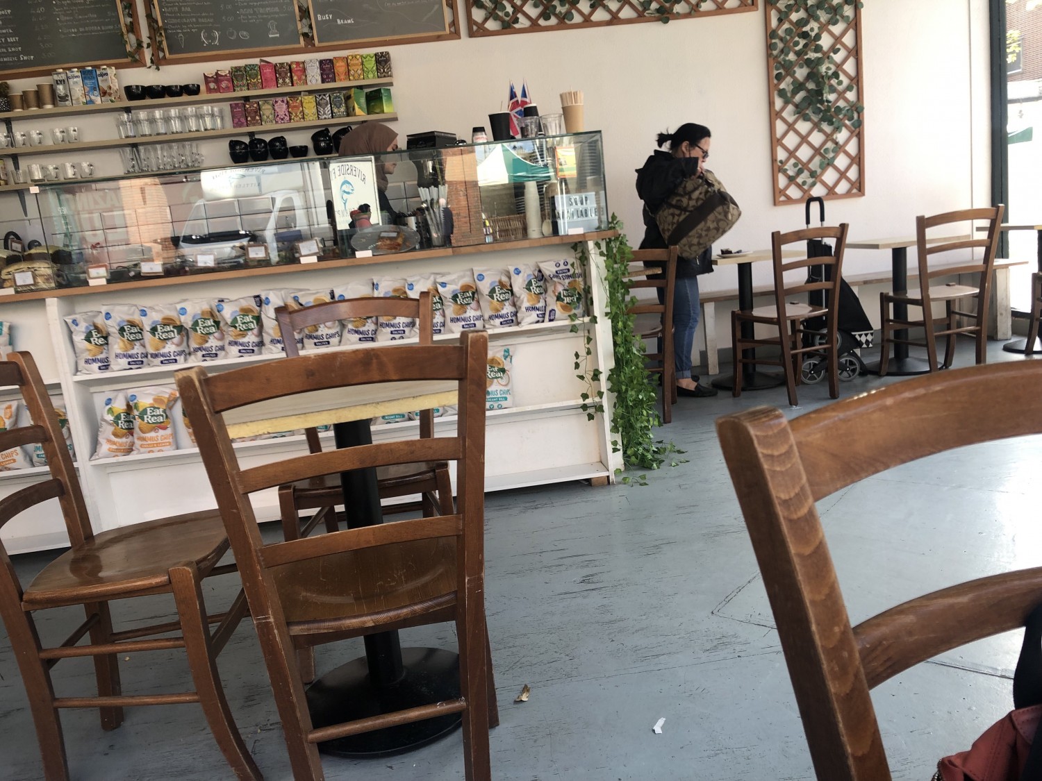 Serving counter, tables and chairs in cafe. Counter is off white and there is a glass screen with food displayed behind it.