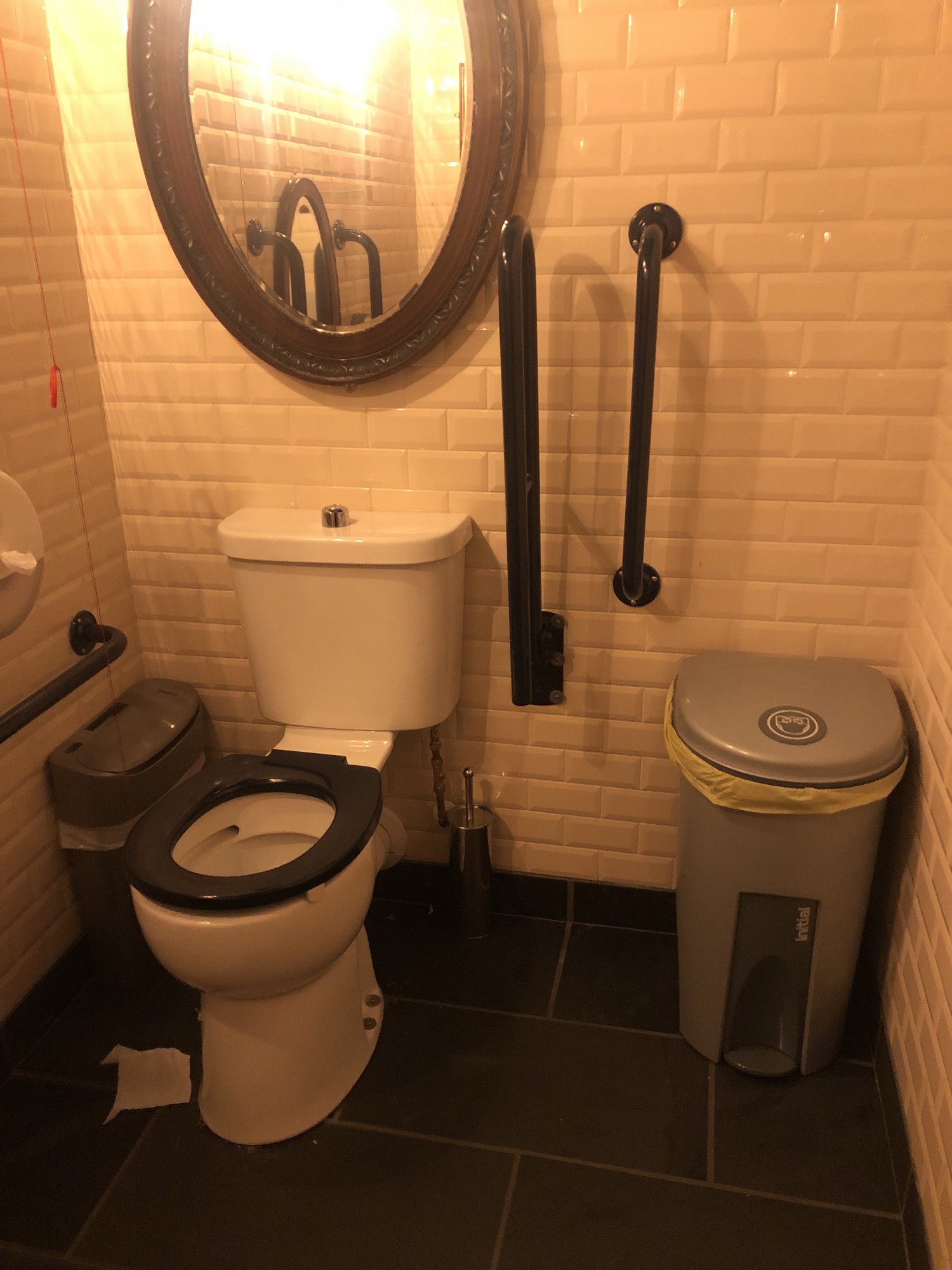 White tiled walls, with (from left to right) black grab rail, dark grey sanitary bin, white bathroom with black seat, black fold down grab rail and black fixed vertical grab rail, and grey rubbish bin.
