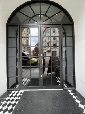 Main entrance. Glass double door with black panelling. Handle in the middle. Pull to open.